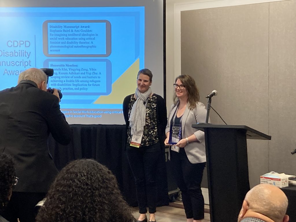 Ami is standing with Dr. Claudia Sellmaier accepted the CSWE Disability Manuscript Award. They are located at the front of the room in front of a screen..
