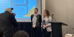 Ami is standing with Dr. Claudia Sellmaier accepting the CSWE Disability Manuscript Award. They are located at the front of the room in front of a screen. Ami is wearing a grey blazer and holding the award.