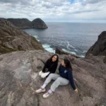 Mariela (left) and Sara (right) sitting together on a rock in a hike. The ocean is in the background.