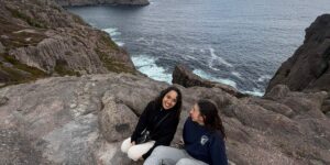 Mariela (left) and Sara (right) sitting together on a rock in a hike. The ocean is in the background.